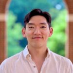 Headshot of Sangkyun Cho. He has short dark hair, brown eyes, and light skin-tone. He is wearing a white button down dress shirt and is standing outside in brick archway with trees in the background.