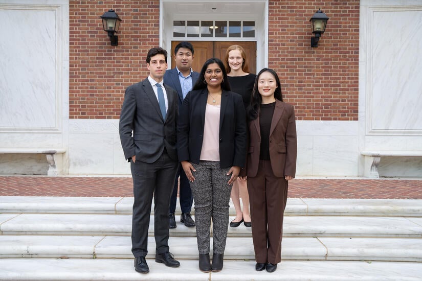 group photo of five students; three women and two men. They are wearing business casual clothes and standing on marble steps in front of a red brick and marble building.