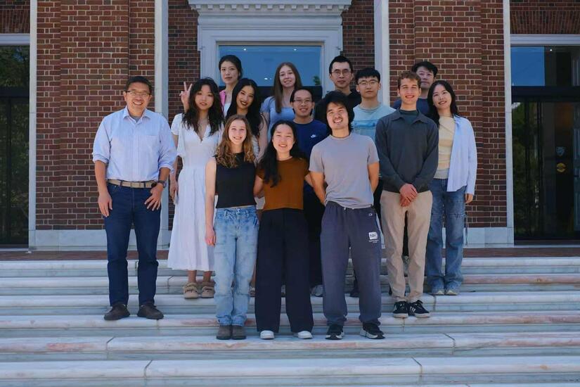 Group photo of 14 adults on marble steps in front of a red brick building. 