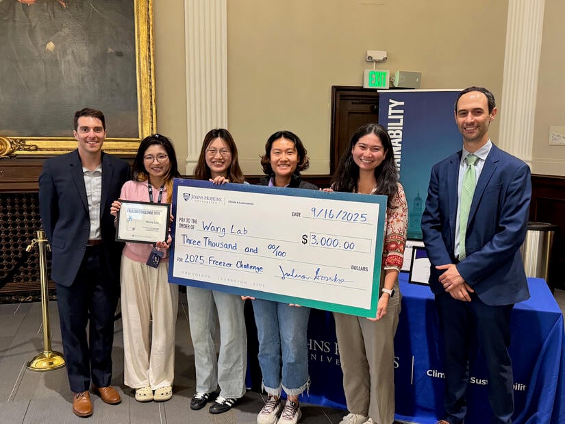 Group photo six people inside a building. The four people in the middle are holding an oversized check for $3,000.