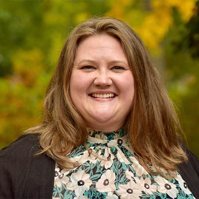 Headshot of a women. She is wearing a white and green floral shirt with a black cardigan. She has shoulder length blonde hair, blue eyes, and light skin tone. She is standing outside and a tree with green and yellow leaves are in the background.