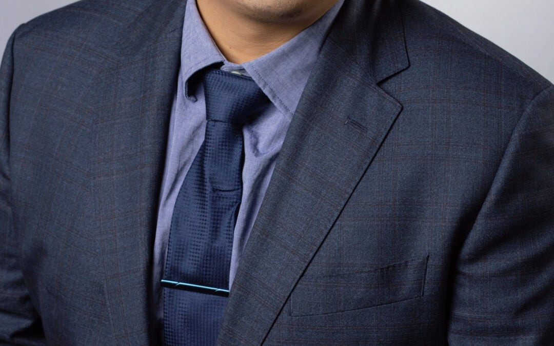 Headshot of a man in a dark blue suite jacket, tie, and dress shirt. He has medium-light skin tone, almond shaped eyes, dark brown eye color and short black hair.