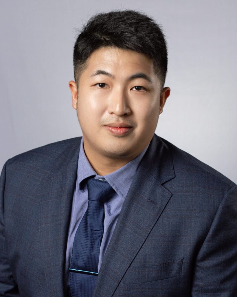 Headshot of a man in a dark blue suite jacket, tie, and dress shirt. He has medium-light skin tone, almond shaped eyes, dark brown eye color and short black hair.