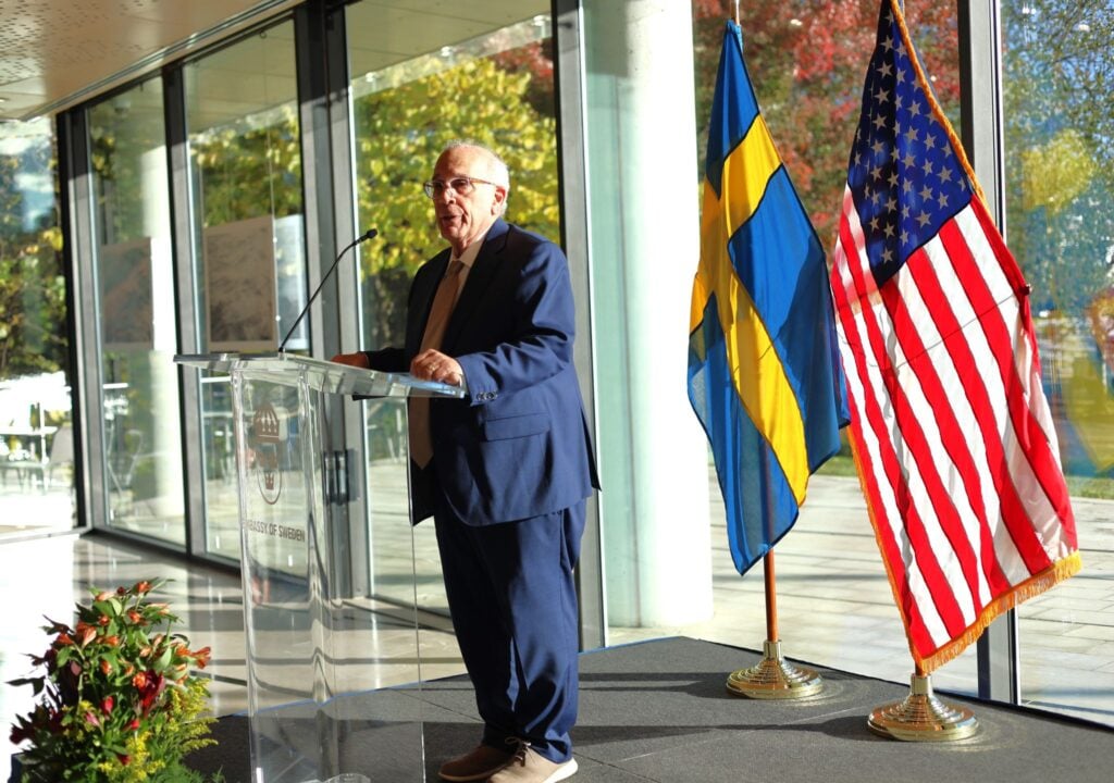Man in blue suite standing in front of a giving a presentation. He is standing in front of a clear podium with the flats of the United States of America and Sweden behind him.