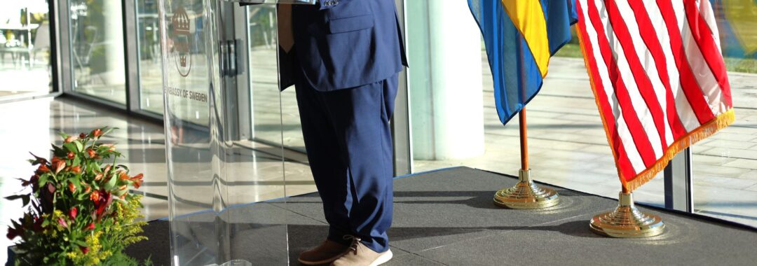 Man in blue suite standing in front of a giving a presentation. He is standing in front of a clear podium with the flats of the United States of America and Sweden behind him.