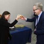 Women on left in black business suit and man on right in blue business suite tap drinking glasses to celebrate an award.